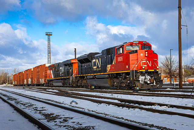 Red CN locomotive 8835 leads CN E271 past orange freight cars on snow-covered tracks at Markham Yard, Homewood, Illinois, with a blue sky and yard infrastructure in the background.