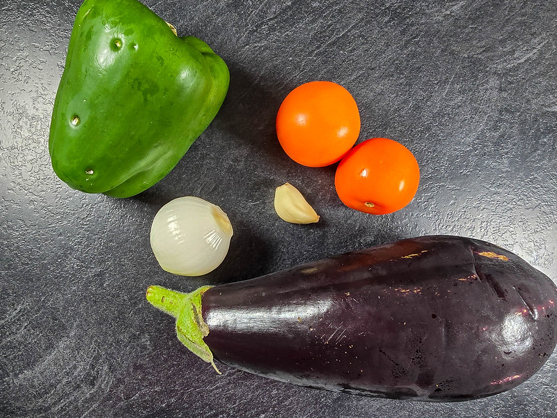 A few vegetables on a black kitchen worktop: an eggplant, two tomatoes, a garlic clove, one onion, one green pepper