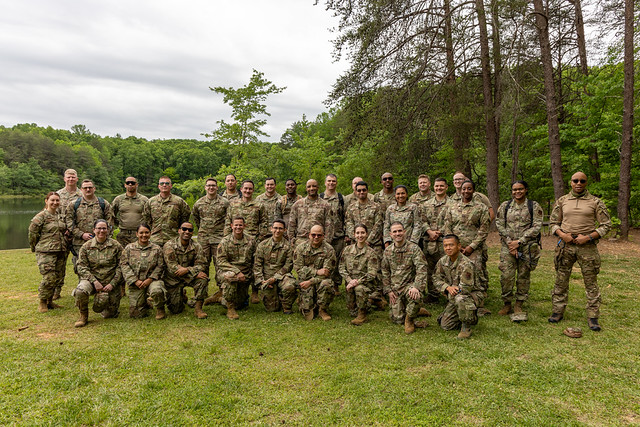 Group of U.S. Airmen in camouflage kneeling and standing on a grassy lakeside at Crowders Mountain State Park, North Carolina, during Expeditionary Skills Training.