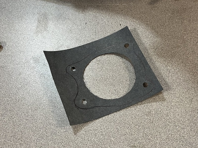 A black gasket made of rubber-like material, featuring a circular hole in the center and two small holes at the edges, placed on a textured surface.