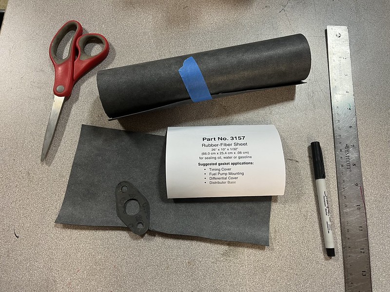 A flat workspace with a pair of red scissors, a roll of rubber-fiber sheet secured with blue tape, a label indicating part number 3157 and gasket applications, a small black rubber piece, a ruler, and a black marker.