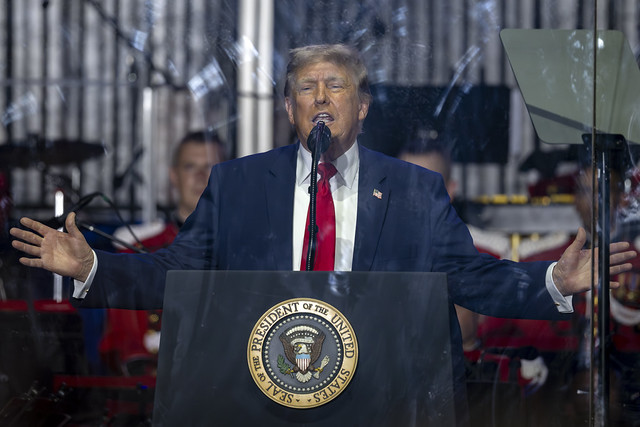 Speaker in a navy suit and red tie at a podium with presidential seal, microphone centered, arms wide, behind a clear protective barrier on an outdoor stage.