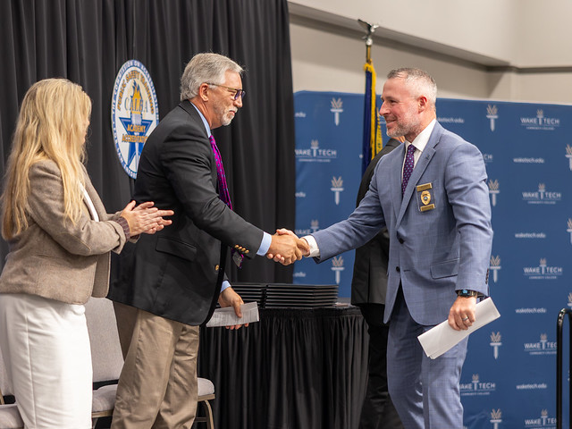 Two men in suits shake hands on a stage during a BLET graduation at Wake Tech; a woman applauds nearby, with diplomas on a table and a blue Wake Tech backdrop.