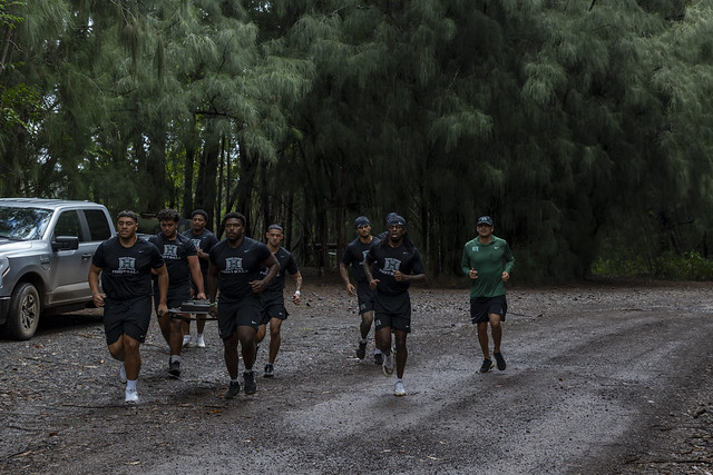 Football team in black shirts sprint along a damp gravel road under a dense pine forest; trainer in green runs beside them; a silver pickup sits to the left.