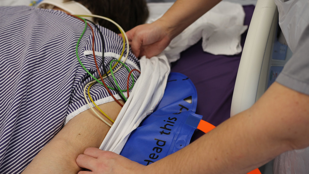 A patient lying prone on a hospital bed with the sheet pulled back to reveal a blue mat-like device beneath.