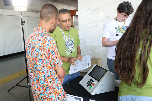 Três pessoas ao redor de uma urna eletrônica sobre uma mesa em ambiente público; um homem verde aponta para um folheto, outro observa com estampa colorida, e o terceiro de camiseta branca olha o material; iluminação suave à esquerda.