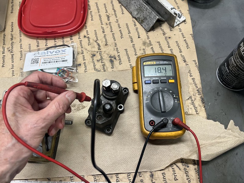 A hand holding a red multimeter probe testing an automotive component, with a digital multimeter displaying a reading of 18.4 volts on a workbench covered with various tools and parts.