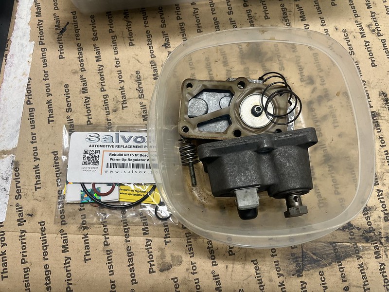 A clear container holding automotive parts including a warm-up regulator, springs, gaskets, and a rebuild kit for Bosch components, placed on a Priority Mail service background.
