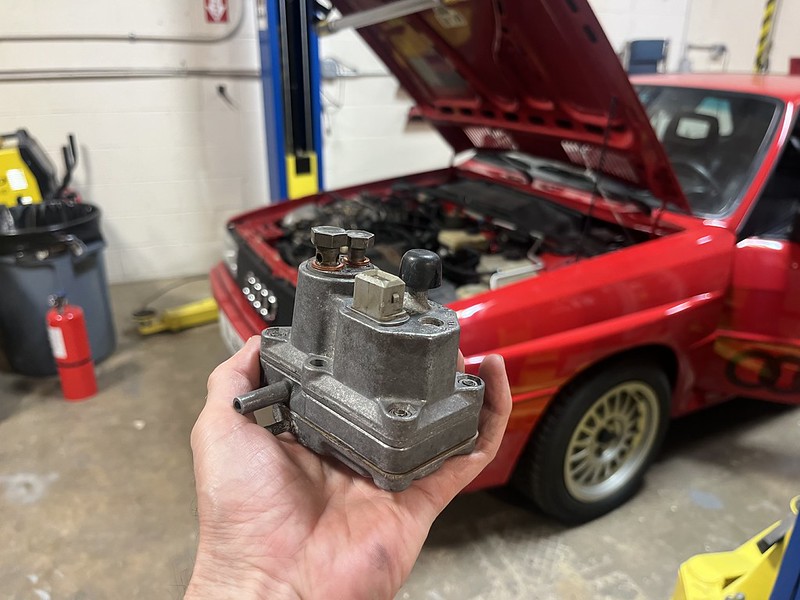 A hand holding a mechanical component, possibly a fuel pressure regulator, in a garage with a red car in the background. The car's hood is open, revealing its engine.