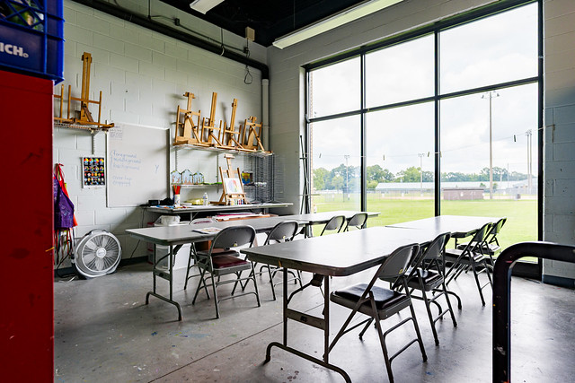 Wide-angle interior of a daytime art classroom before renovations, with long folding tables, metal chairs, whiteboard, shelving with easels, red storage cabinet, floor fan, and large windows overlooking a grassy field.