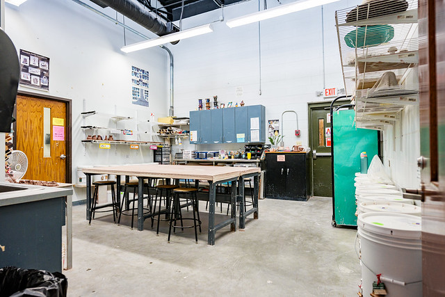 Interior of Jaycee Park Center for Arts & Crafts ceramics studio before renovations: long central worktable with stools, blue wall cabinets, shelves of pottery, sink area, green storage refrigerator, and white buckets along the right, under bright fluorescent lighting.