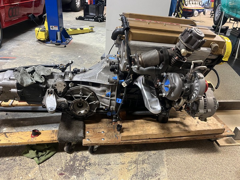 A disassembled engine and transmission on a wooden platform, featuring various mechanical components and tools in a workshop setting.