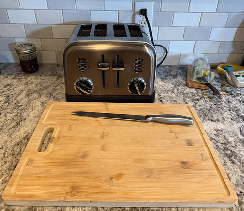 A wooden cutting board with a bread knife in front of a toaster.