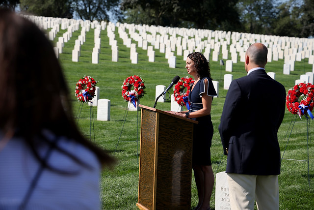 Speaker at a wooden podium delivers remarks during a wreath-laying ceremony at Wood National Cemetery; rows of white headstones line a green slope, with red wreaths and blue ribbons nearby.