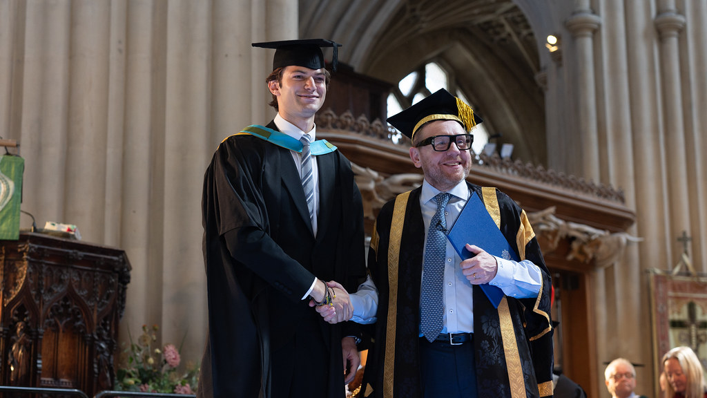 Simon dressed in graduation robes shaking hands with the dean during graduation ceremony.