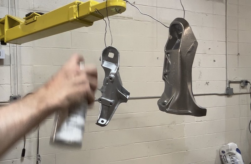 A hand holding a spray can applies paint to metallic car parts hanging from a yellow overhead crane in a workshop.