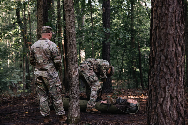 Two soldiers in woodland camouflage perform a casualty-care drill in a pine forest; one secures a combat casualty manikin to a rescue system while the other observes.