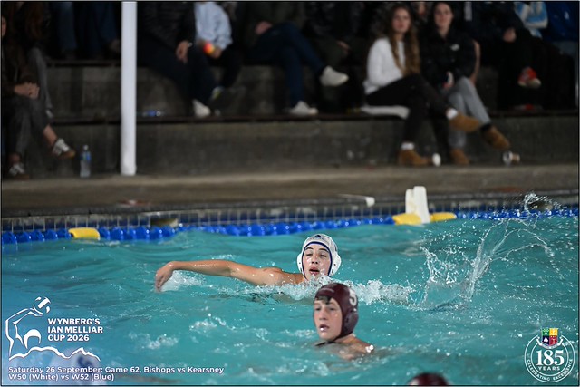 Two water polo players in white and maroon caps sprint across turquoise pool as water sprays; spectators sit in concrete stands behind, with Wynberg Ian Melliar Cup branding bottom-left.