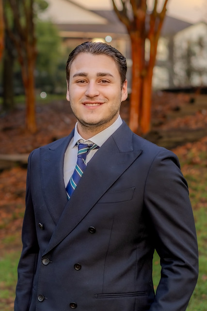 Nick Allen smiles for a portrait outdoors, wearing a navy double-breasted suit and blue striped tie, with trees and a building softly blurred in the background.