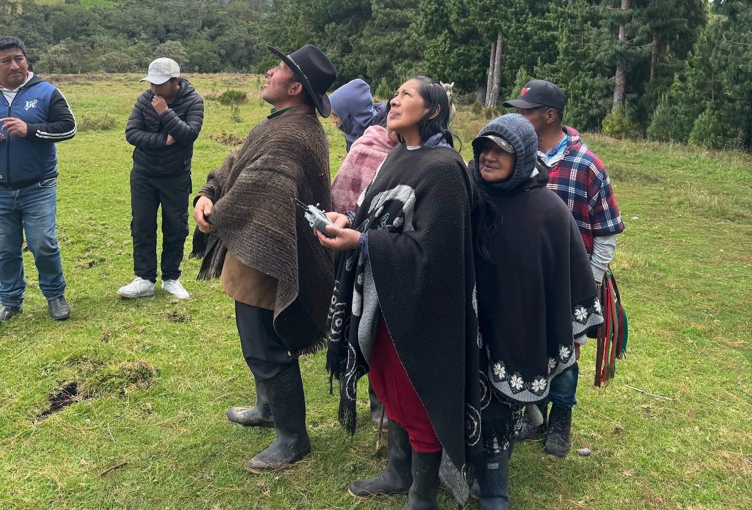 Community members follow a drone flight in a highland meadow while one participant holds the remote controller during an open mapping workshop in Colombia.