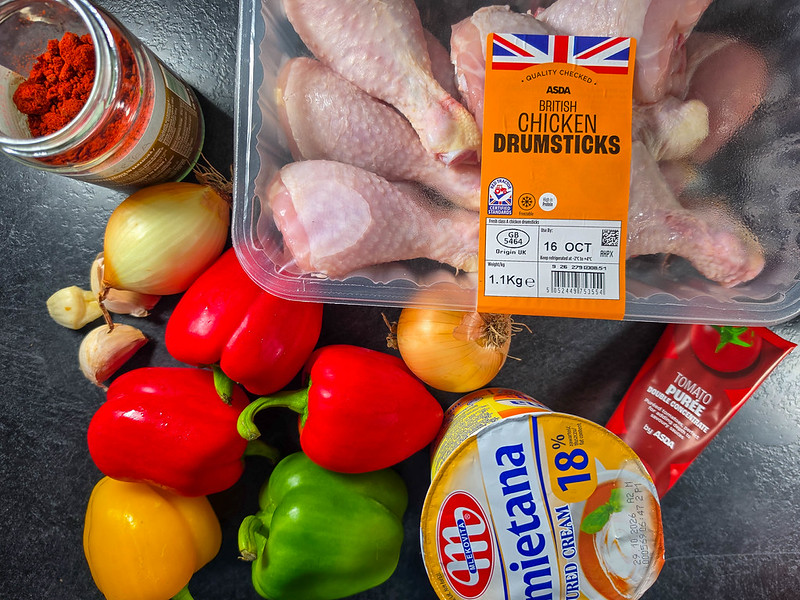 Some of the ingredients for this recipe on a black kitchen countertop: peppers, onions, garlic, chicken drumsticks, tomato puree, a jar with red paprika in it and an orange cup of sour cream