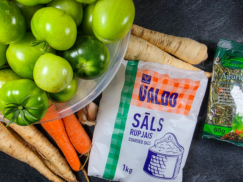 A bowl filled with green tomatoes, a packof coarse salt, carrots, horseradish, garlic, a pack of dried herbs