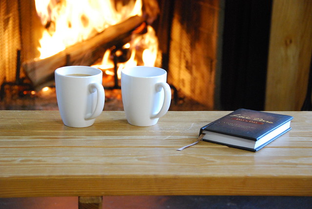 A fireplace roars in the background, two coffee cups and a book sit on a bench in the foreground.