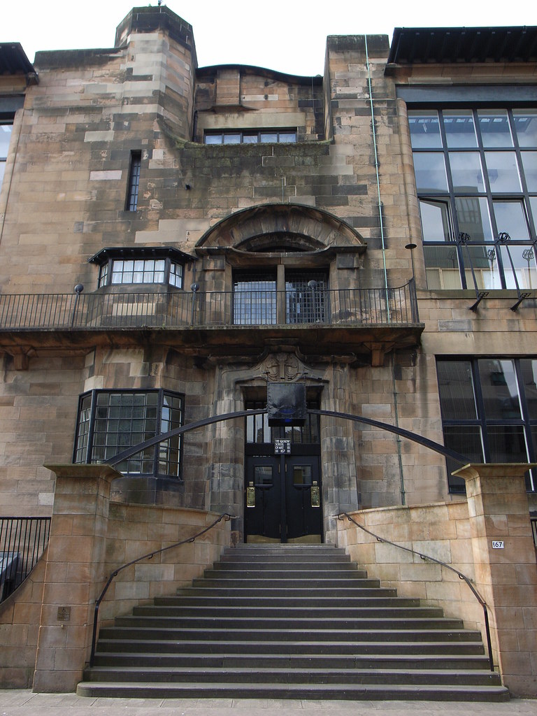Doorway, Glasgow School of Art: A large simple, brick building with a balcony on the second level, a curvy staircase entrance and rectangular windows. 