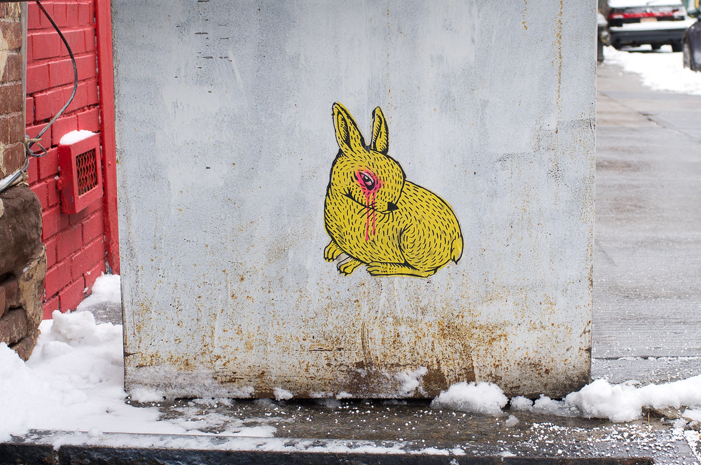 Yellow sticker of a crouching rabbit with a weeping pink eye on a weathered utility box in the snow.