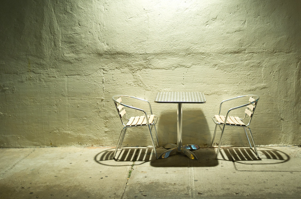 Two outdoor cafe chairs and a small table lit against a textured stucco wall at night in New York City.