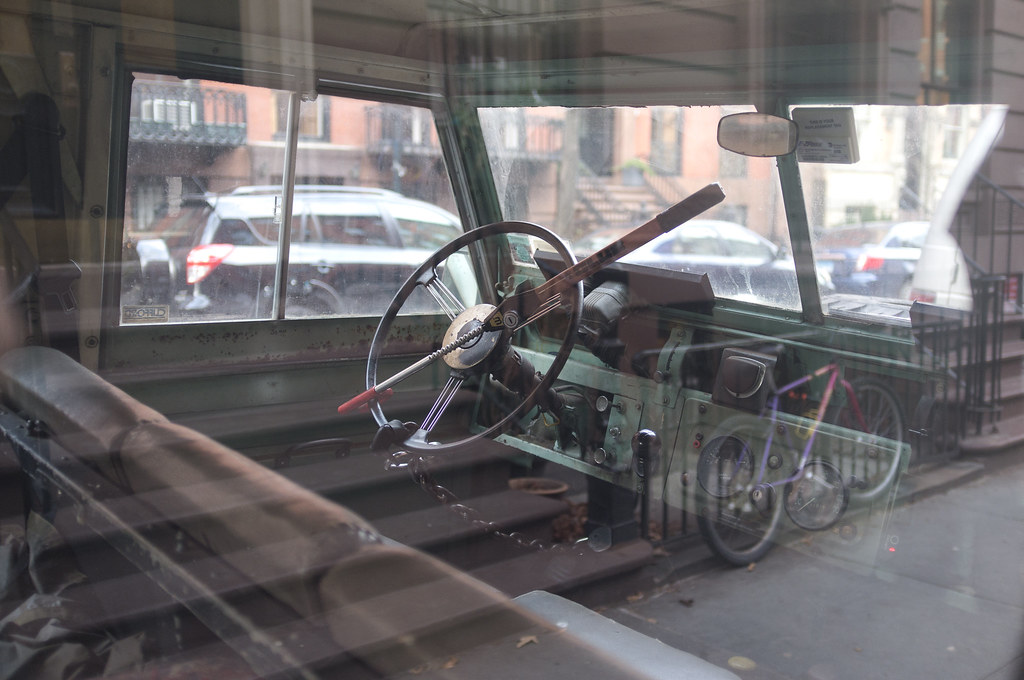 View into the spartan cabin and dashboard of a vintage Land Rover parked on a brownstone street.