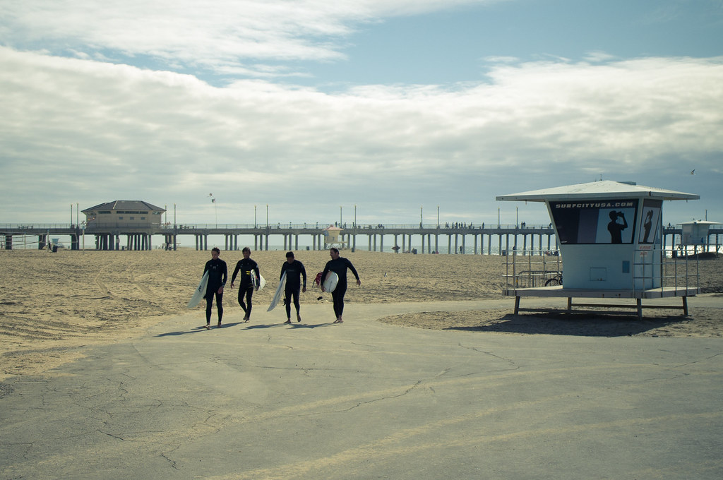 Surfers in wetsuits carrying boards along a path toward the Surf City USA tower and Huntington Beach Pier.