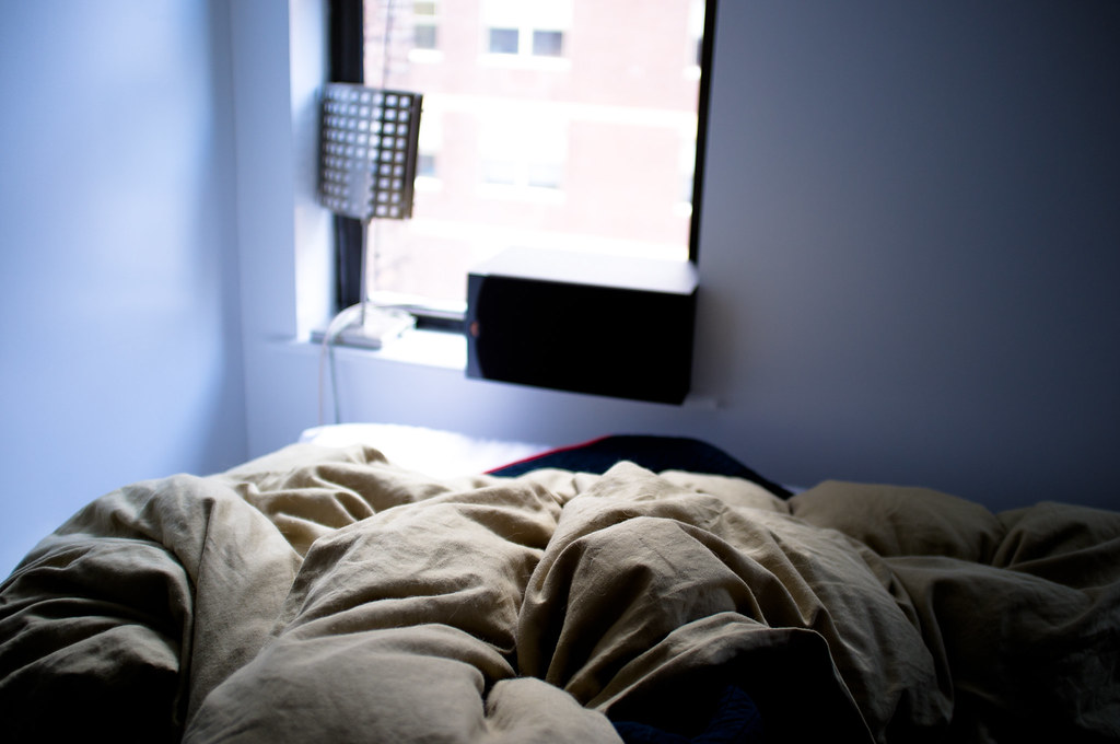 Morning light over rumpled bed covers looking toward a window sill with lamp and speaker.