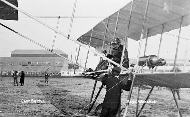 Farnborough - Captain C J Burke sat in the cockpit of the pusher biplane that he designed talking to a man stood on the ground. One of the airship sheds can be seen in the background.