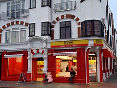 The ground floor of the above-pictured premises, now painted in bright yellow and red and with a sign reading “Lobo Fresh Halal Meat”.  A butchers counter is visible inside.  One of the windows appears to lack glazing, and a person is sitting on the sill with their legs crossed.  The previously blocked-off arched doorway between the two sides has been reopened.