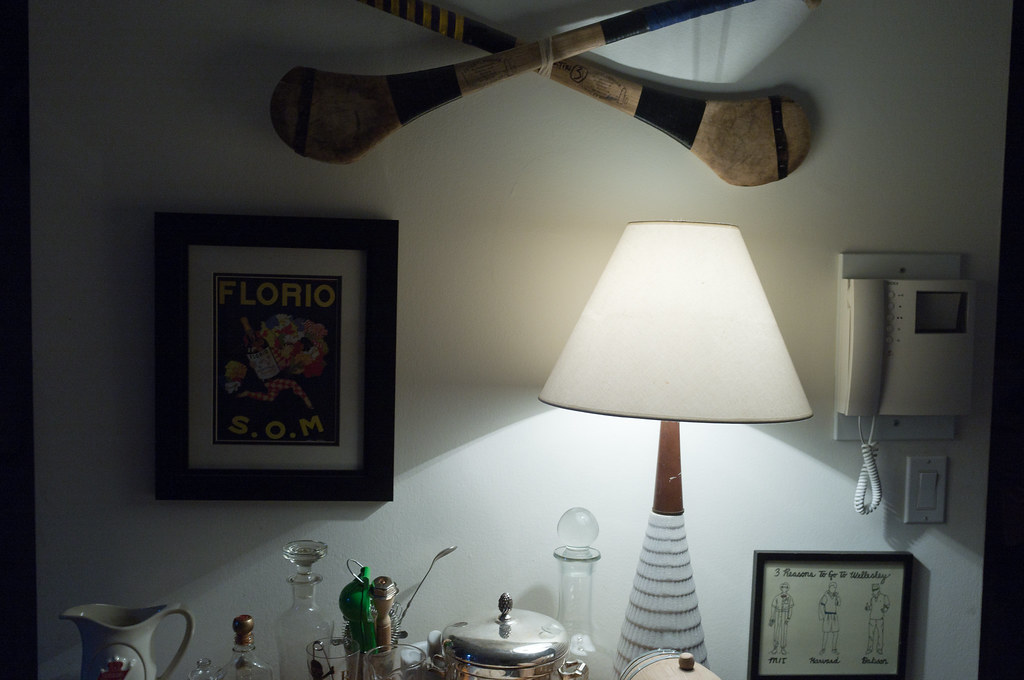 Bar cart with a glowing lamp, decanters, shaker, and crossed vintage wooden hurling sticks on the wall.