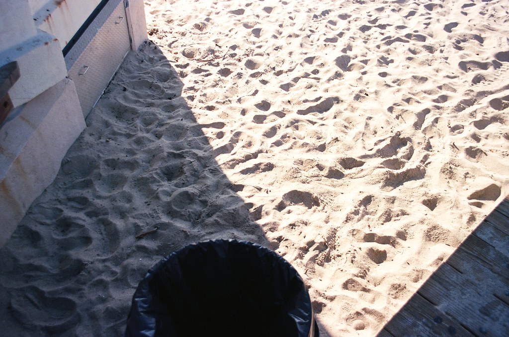 High-angle view of sunlit California beach sand covered in footprints beside a boardwalk and concrete wall.