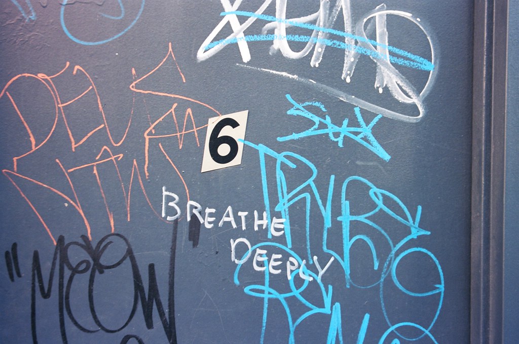 Blue door covered in street art tags and graffiti with handwritten text reading 'BREATHE DEEPLY'.