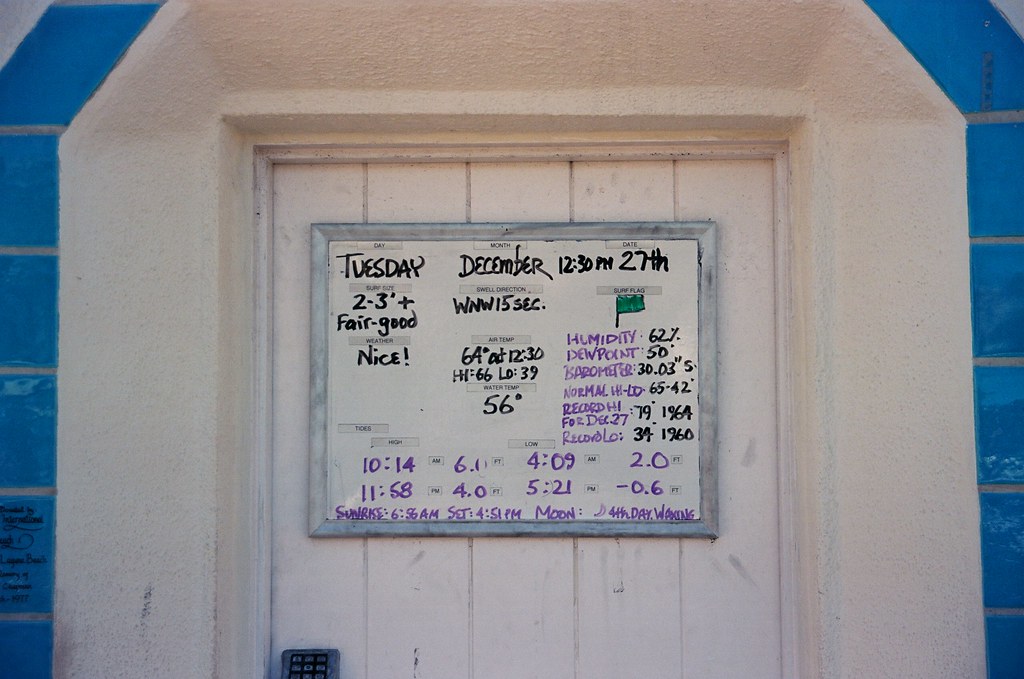 Handwritten whiteboard surf and weather report posted on a wooden door framed by blue tile.