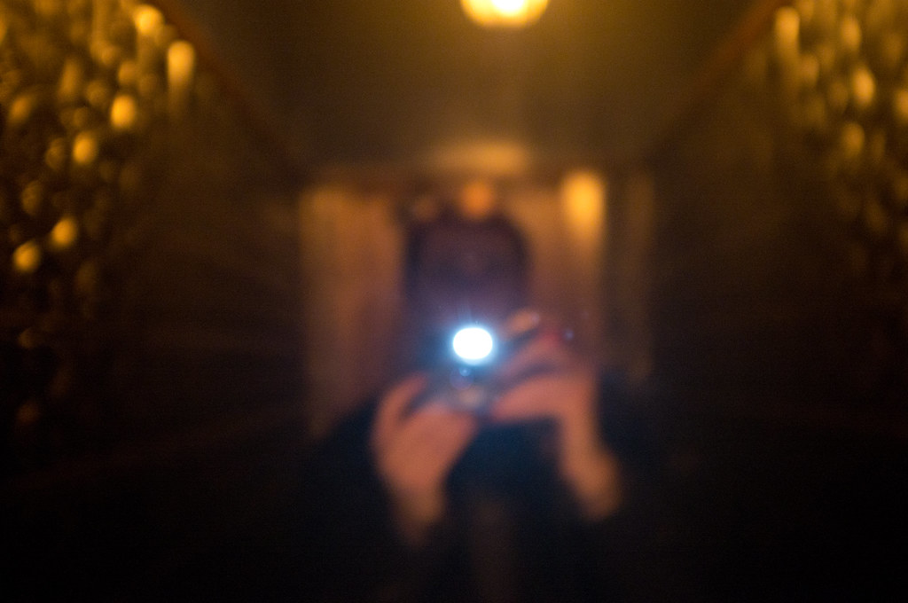 Abstract out-of-focus mirror self-portrait with glowing camera flash in a warm amber-lit hallway.