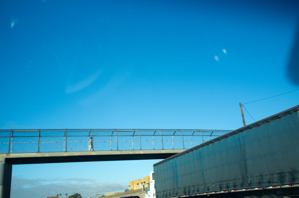 Person crossing a wire-mesh enclosed pedestrian bridge over a highway against a blue California sky.