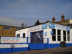 A rectangular brick building painted in white and blue, with a large shuttered car port leading on to an open yard.  A blue gate to the yard closes it off from the road.  Signs on the building and the wall of the yard read “Molbee Motors / Motorists Discount Centre”.