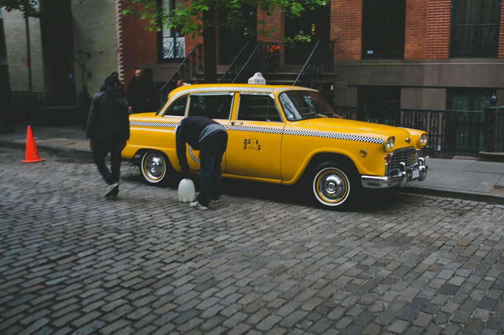 Yellow Checker Marathon taxi cab on a cobblestone street before brownstones as a man tends to the wheels.