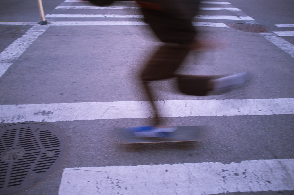 Motion-blurred skateboarder riding quickly across a white painted pedestrian crosswalk on a city street.