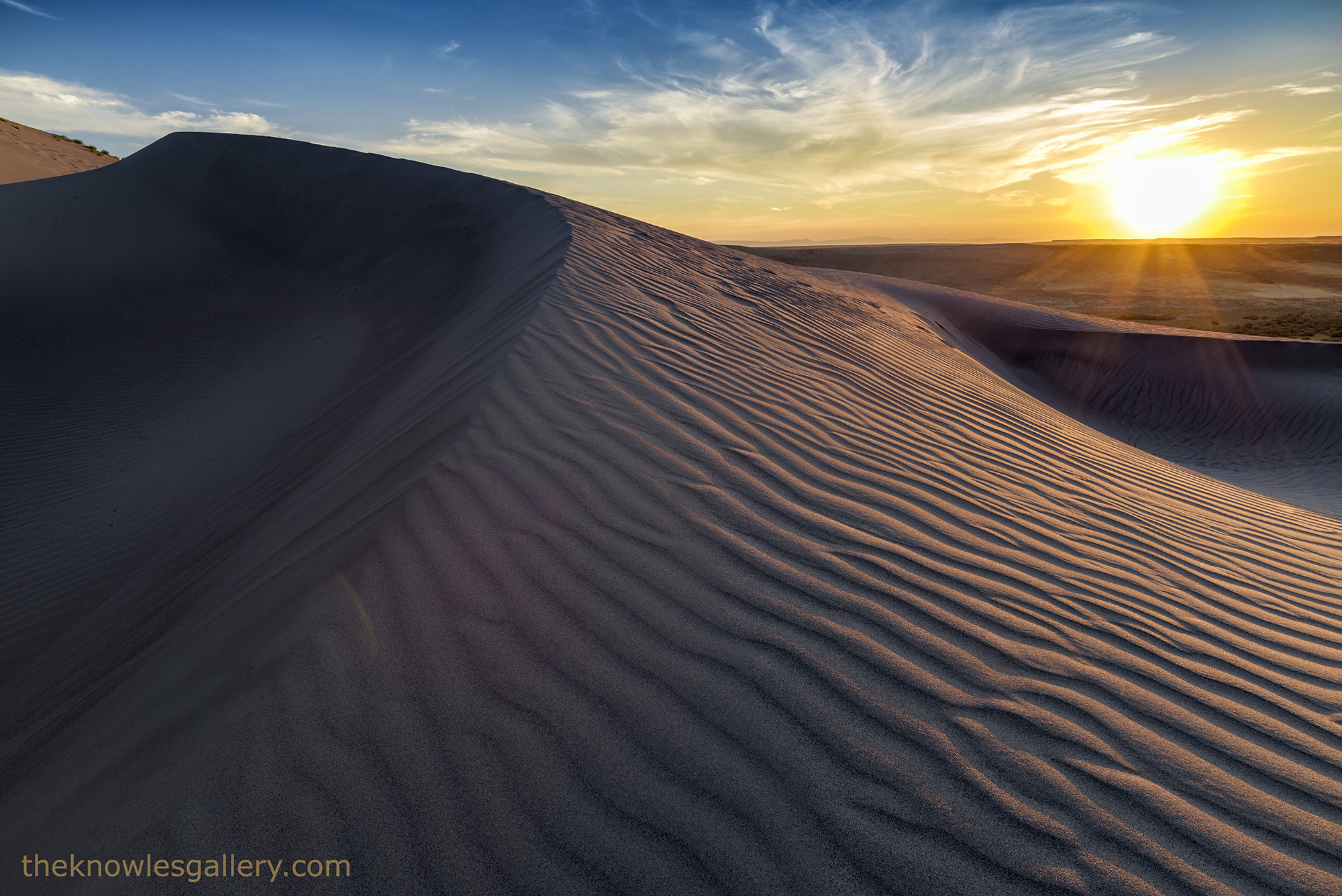 Wind-rippled sand dune at Bruneau Dunes State Park glowing at sunset with a sunburst on the horizon