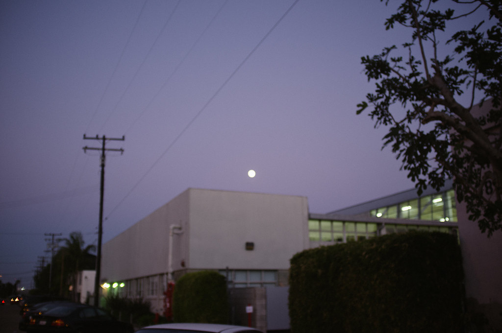 Glowing full moon rising in a twilight sky above a modern commercial building roofline and utility pole.