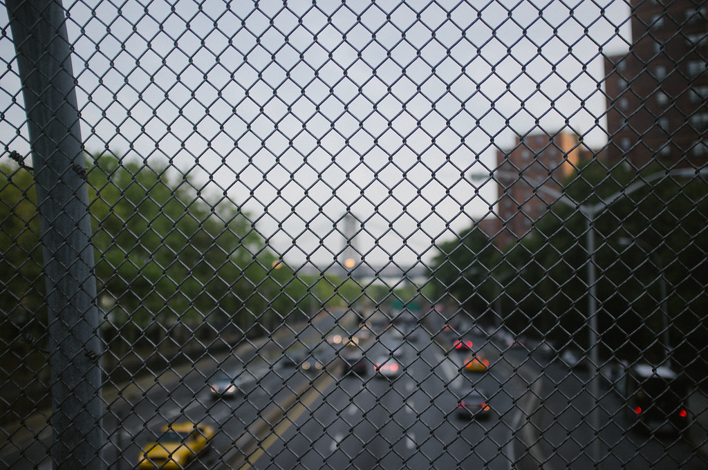 Evening highway traffic on the FDR Drive with blurred headlights seen through a chain-link fence in NYC.