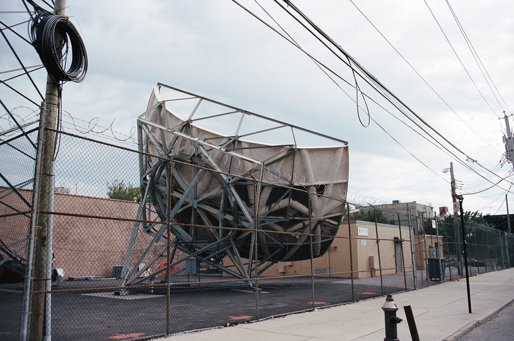 Large curved satellite dish and steel support scaffolding behind a chain-link fence on a city street.