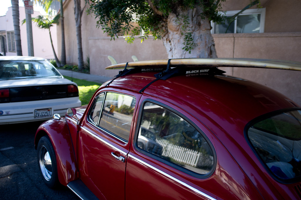Vintage maroon Volkswagen Beetle with a surfboard on its roof racks parked on a sunny California street.