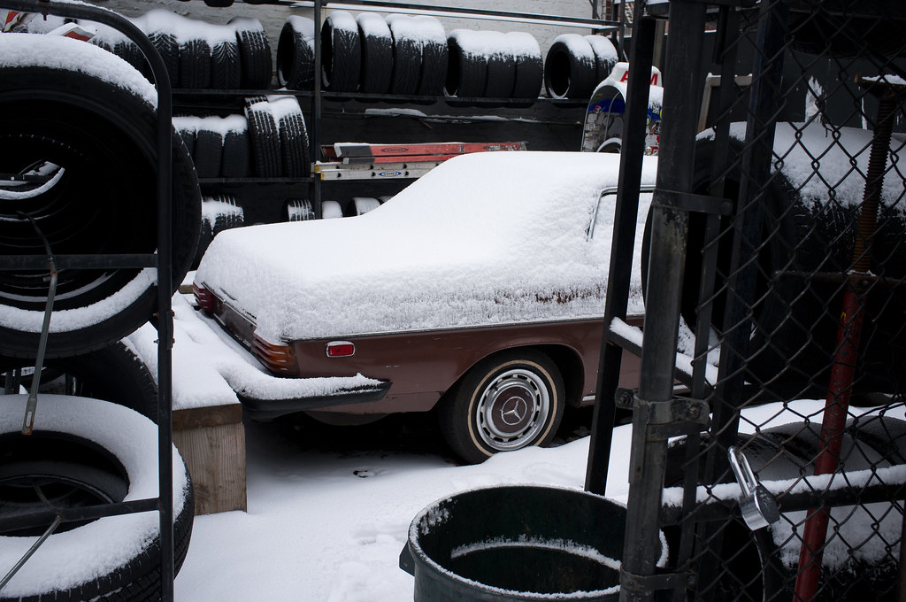Classic brown Mercedes-Benz sedan covered in fresh snow in an outdoor tire shop lot beside stacks of tires.